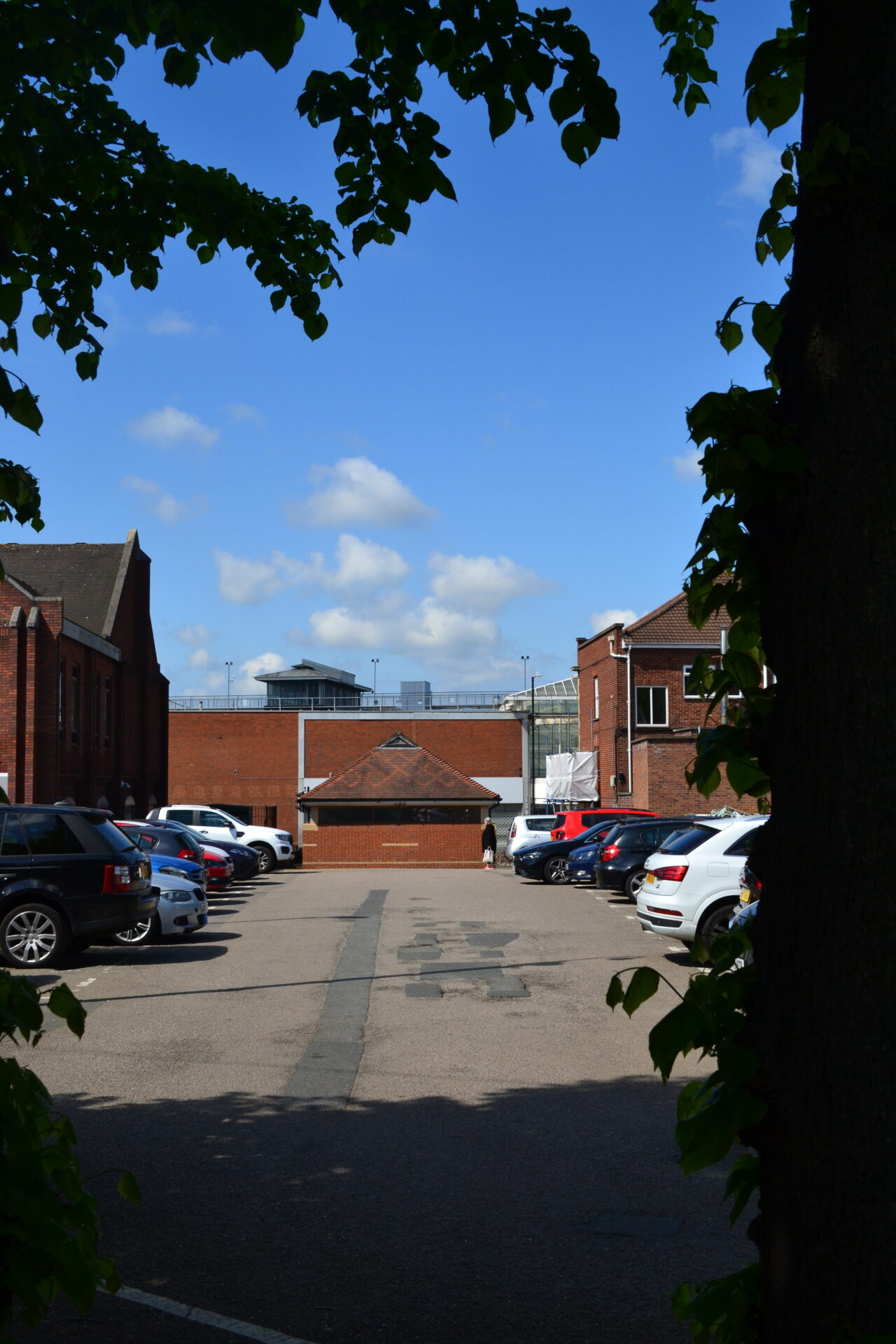 A town centre car park on a sunny day, framed by trees, with rows of parked cars between red-brick buildings and the back of a shopping centre beyond.
