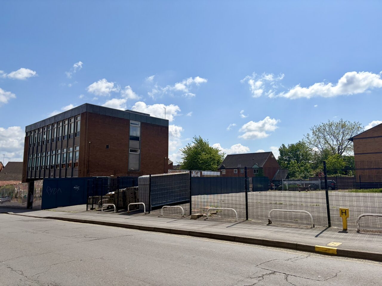 An empty, fenced-off plot beside a three-storey brick and glass office building, with houses and trees behind it, seen from across the road on a sunny day.