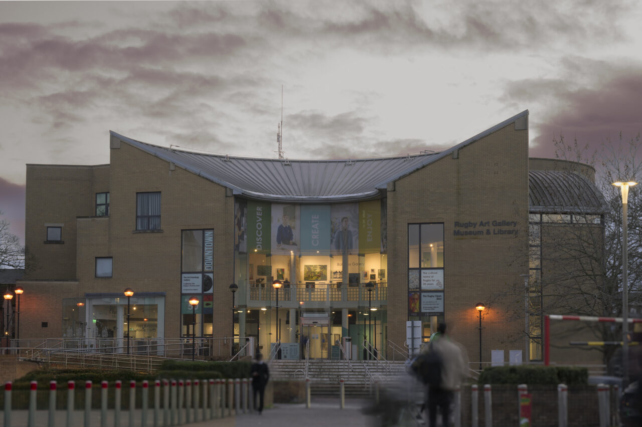 Rugby Art Gallery, Museum and Library at dusk: a buff-brick building with a curved metal roof and a tall glazed centre showing exhibition banners and lit galleries, with steps up to the entrance and people walking past.