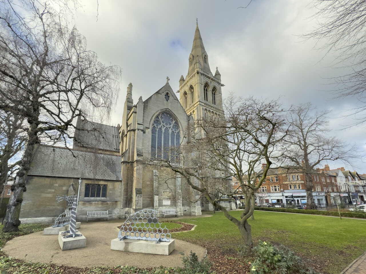 St Andrew's Church on a winter day: a pale stone tower and spire and a large traceried window rise behind bare trees, with silver honeycomb sculptures on a paved circle and a lawn running to the shops on the street beyond.