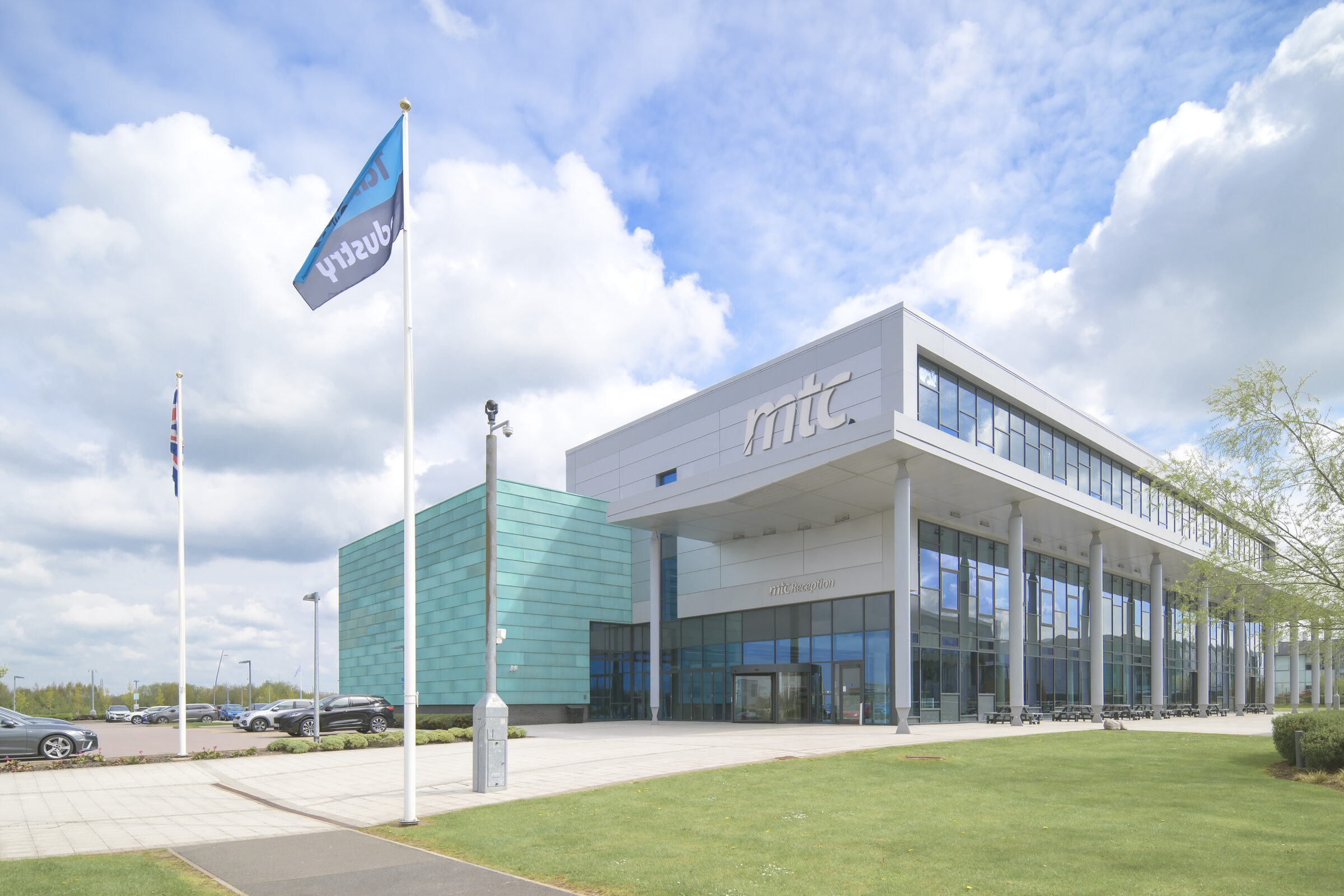The MTC building at Ansty Park: a glass and pale grey office block with a tall colonnade and a turquoise-clad wing, with flags flying out front under a bright, cloudy sky.