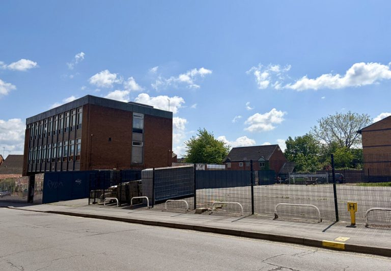 A vacant site on Albert Street behind fencing, with a 1970s office block beyond.