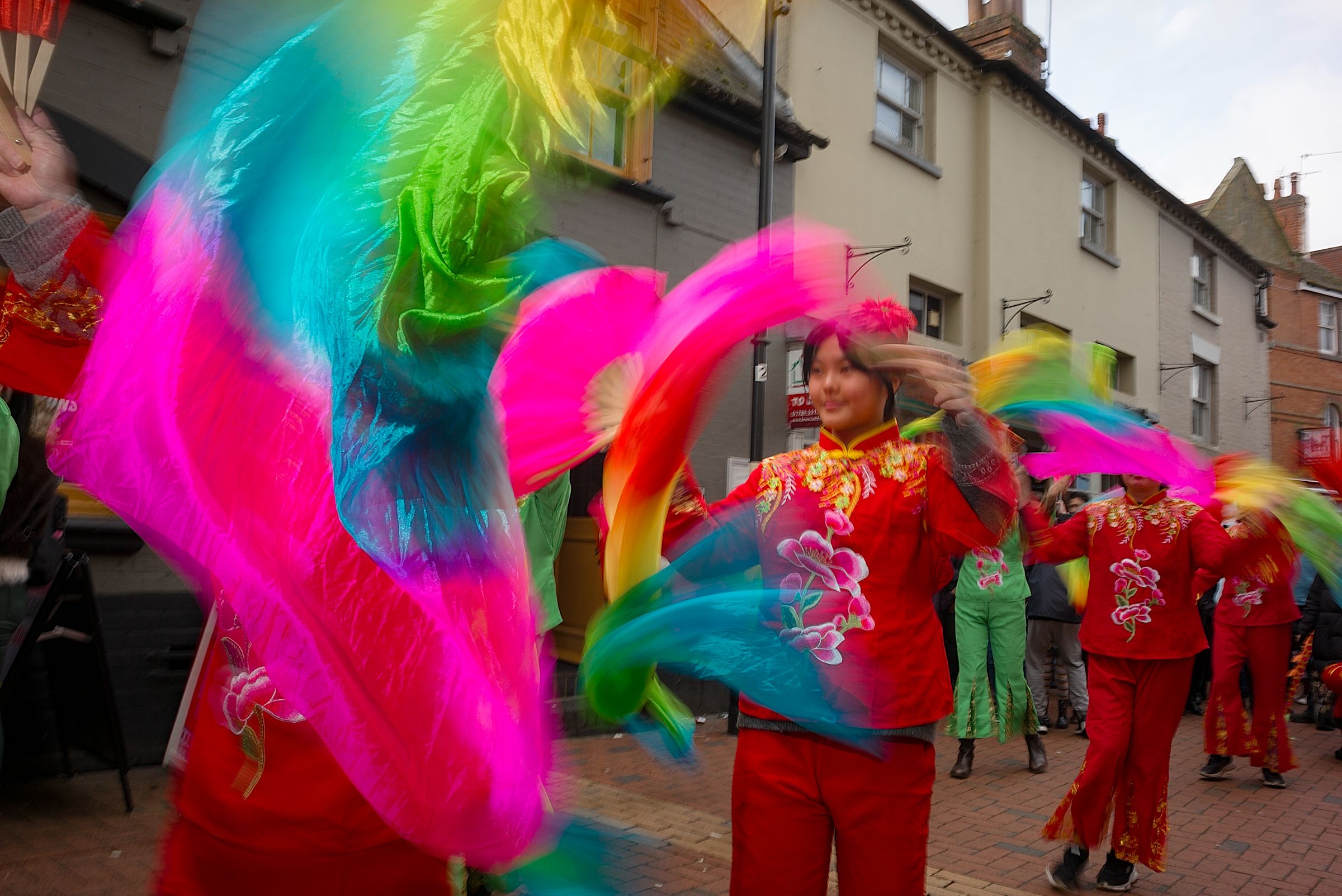 Dancers in red embroidered costumes swirl bright pink, blue and green silk ribbons in a parade along a town centre street.