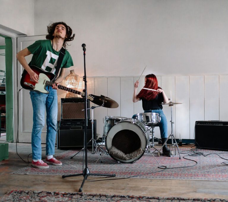 Two young musicians playing guitar and drums in a rehearsal room.