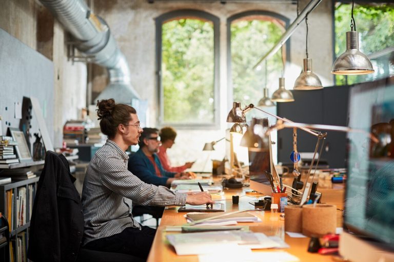 People working at desks in a shared creative workshop with large windows.