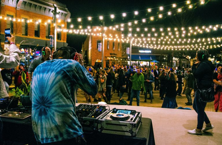 A DJ playing to a crowd at an evening street event under festoon lights.