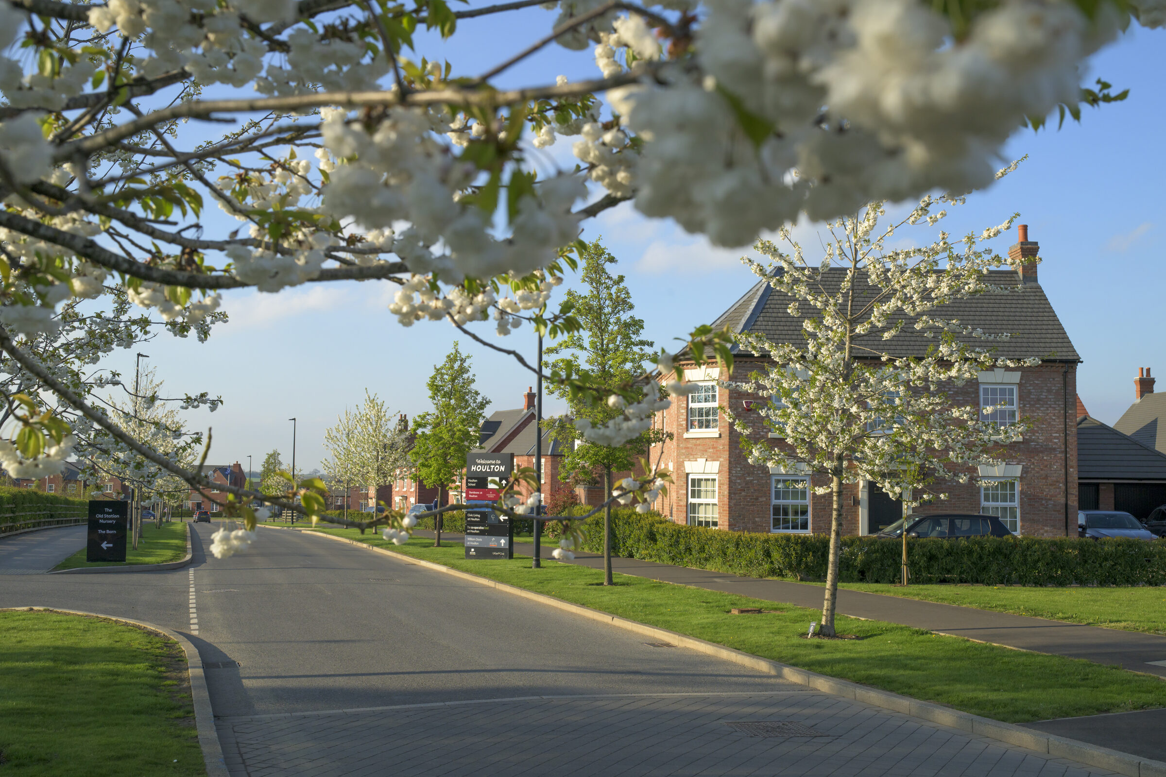 White blossom frames a tree-lined road at Houlton, with new red-brick houses, wide grass verges and a "Welcome to Houlton" sign.
