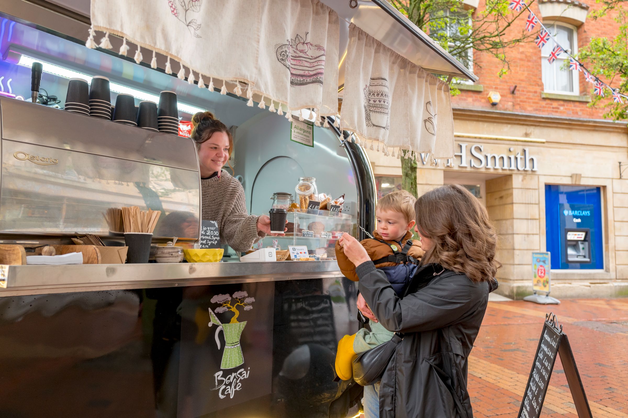 A smiling stallholder hands a coffee from a café trailer to a woman holding a young child, on a pedestrianised shopping street.