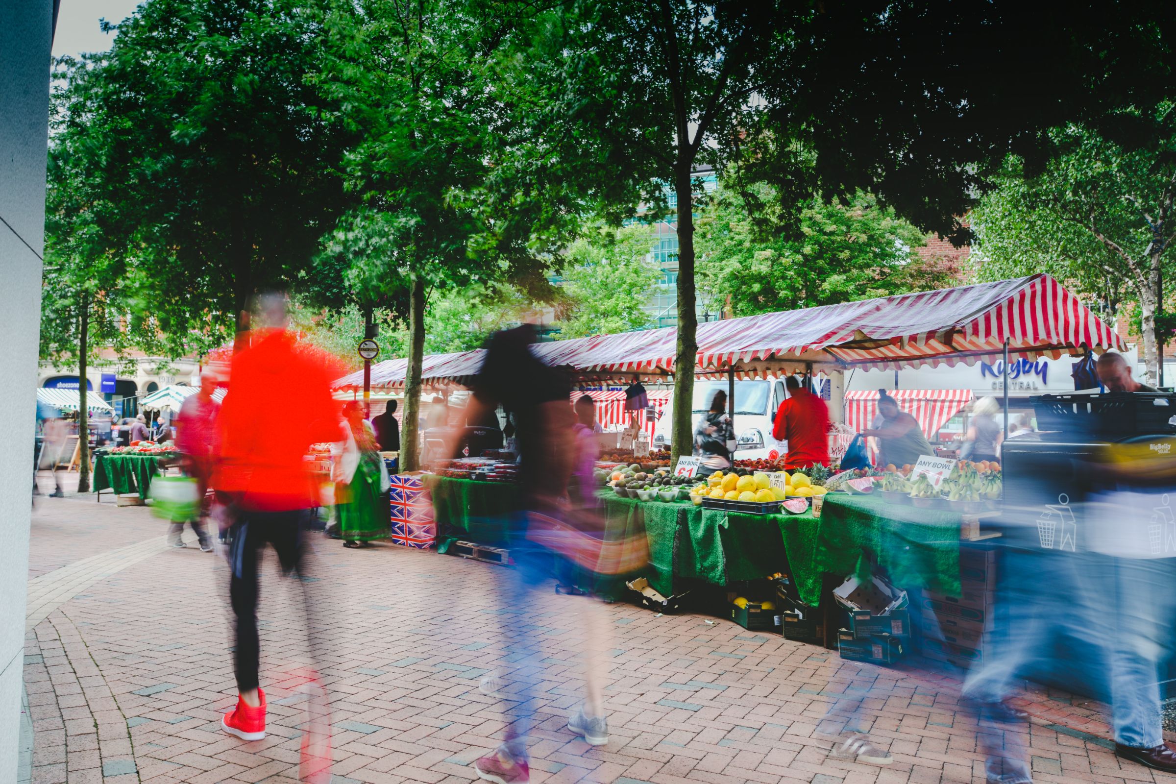 Shoppers walking past fruit and vegetable stalls with red and white striped awnings, under trees on a paved town centre street.