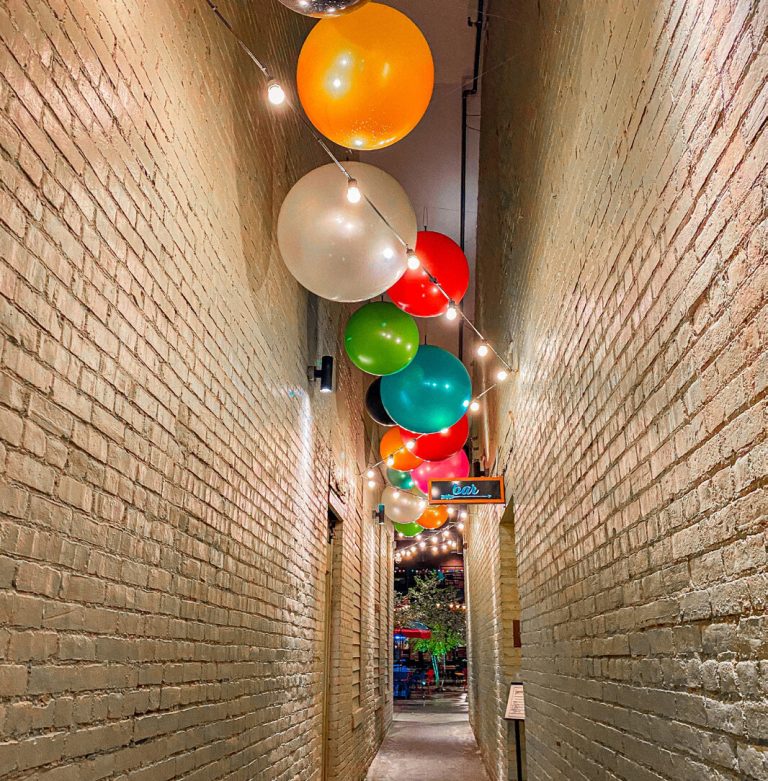 A narrow brick alleyway decorated with hanging coloured balloons.