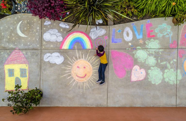 A child drawing on a wall painted with a rainbow, a sun, hearts and the word Love.