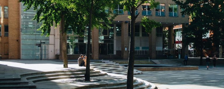 A public square with wide steps, young trees and a modern building behind.