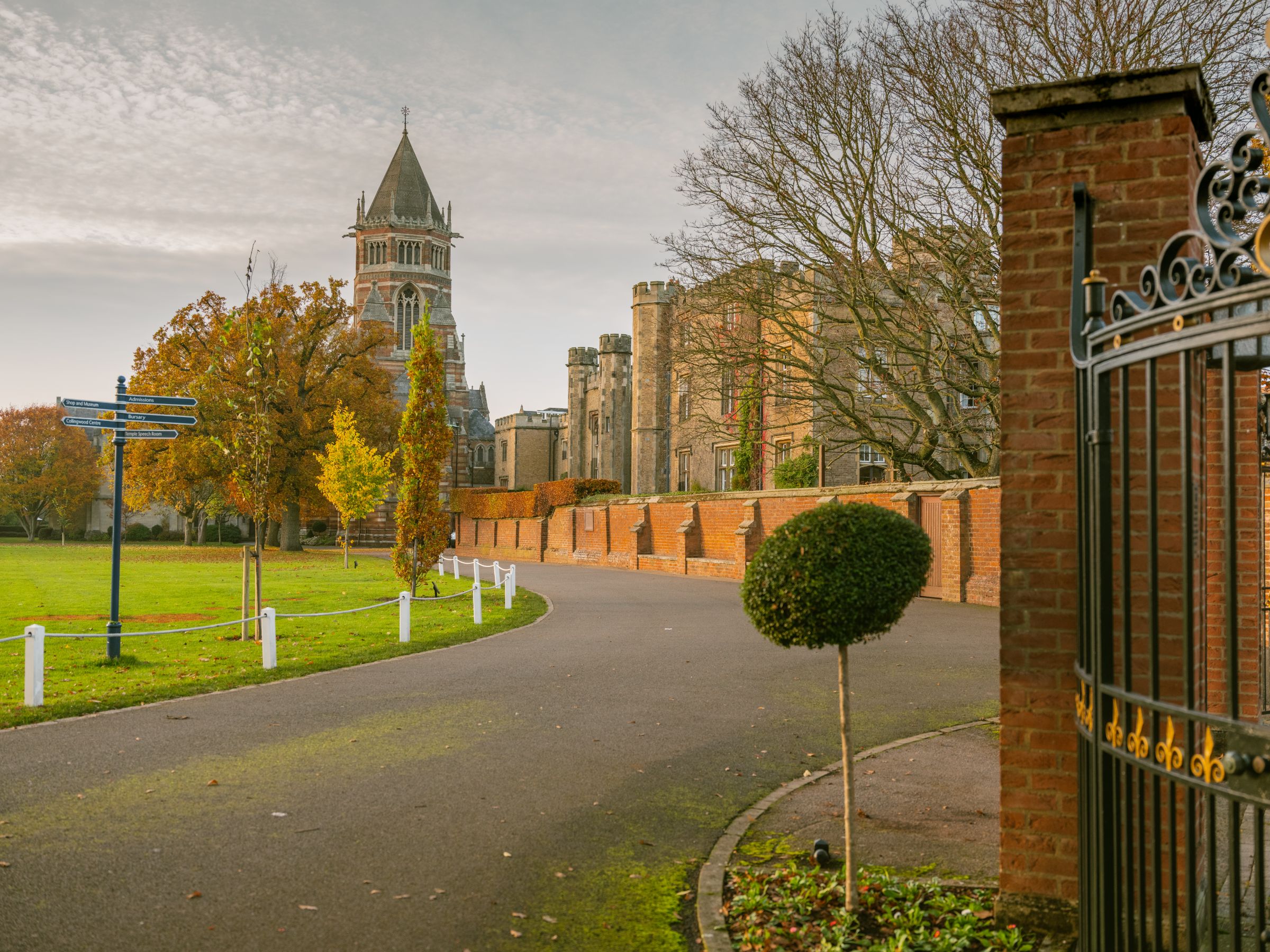 The striped brick tower of Rugby School Chapel and the school buildings beyond a curving drive, with autumn trees and a brick wall.
