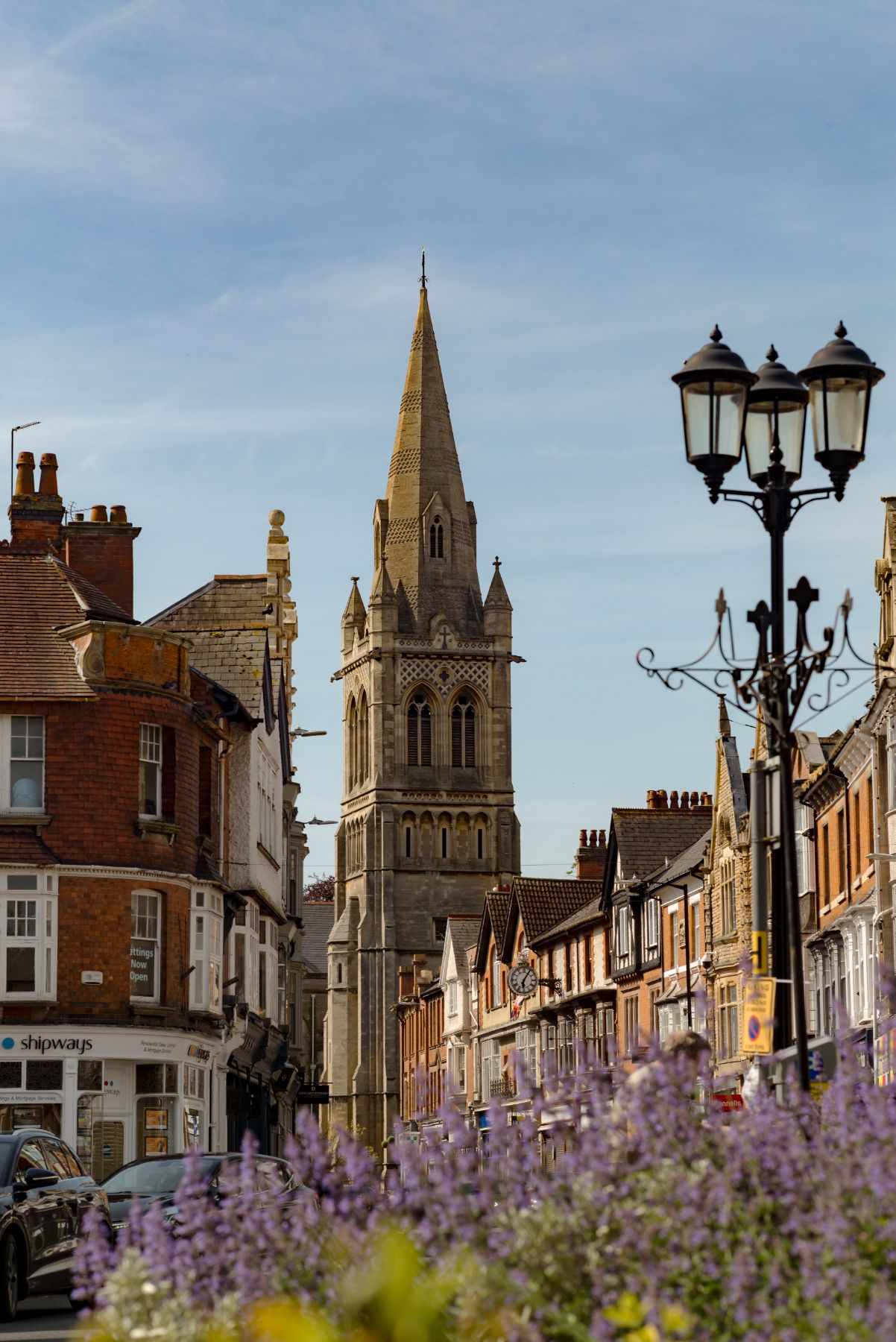 A tall stone church spire rises above Victorian shopfronts and an ornate street lamp, with purple flowers in a planter in the foreground.
