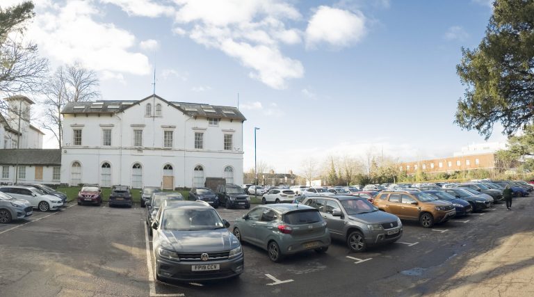 Newbold Road car park, full of cars, with a white listed building beyond.