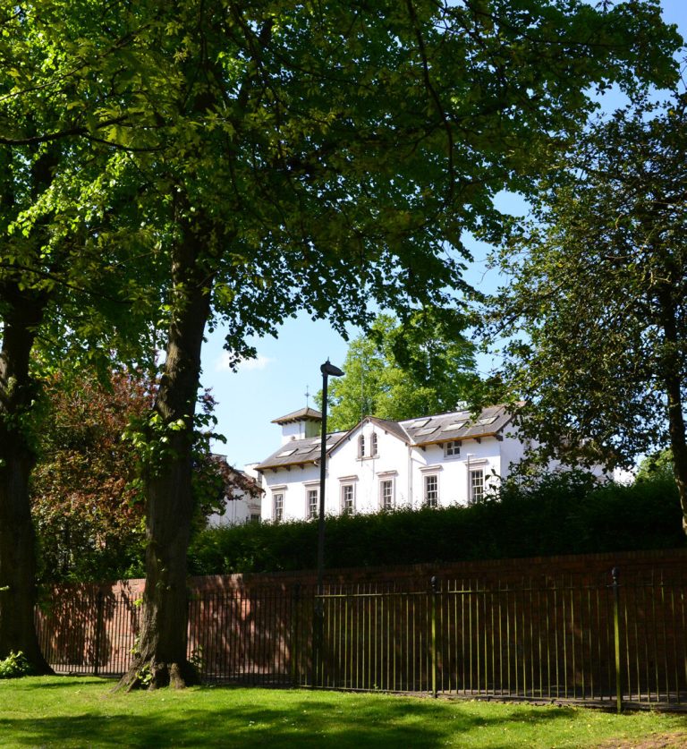 A white listed building glimpsed through large trees from Caldecott Park.