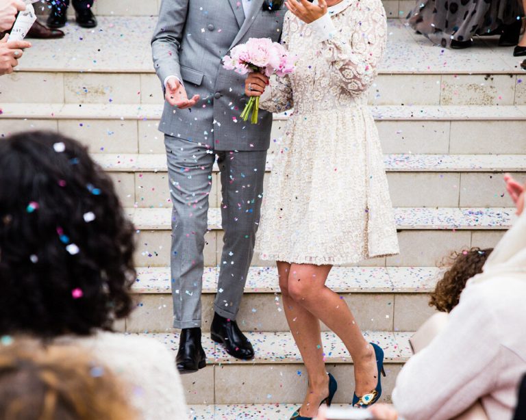 A couple in wedding clothes walking down steps as guests throw confetti.