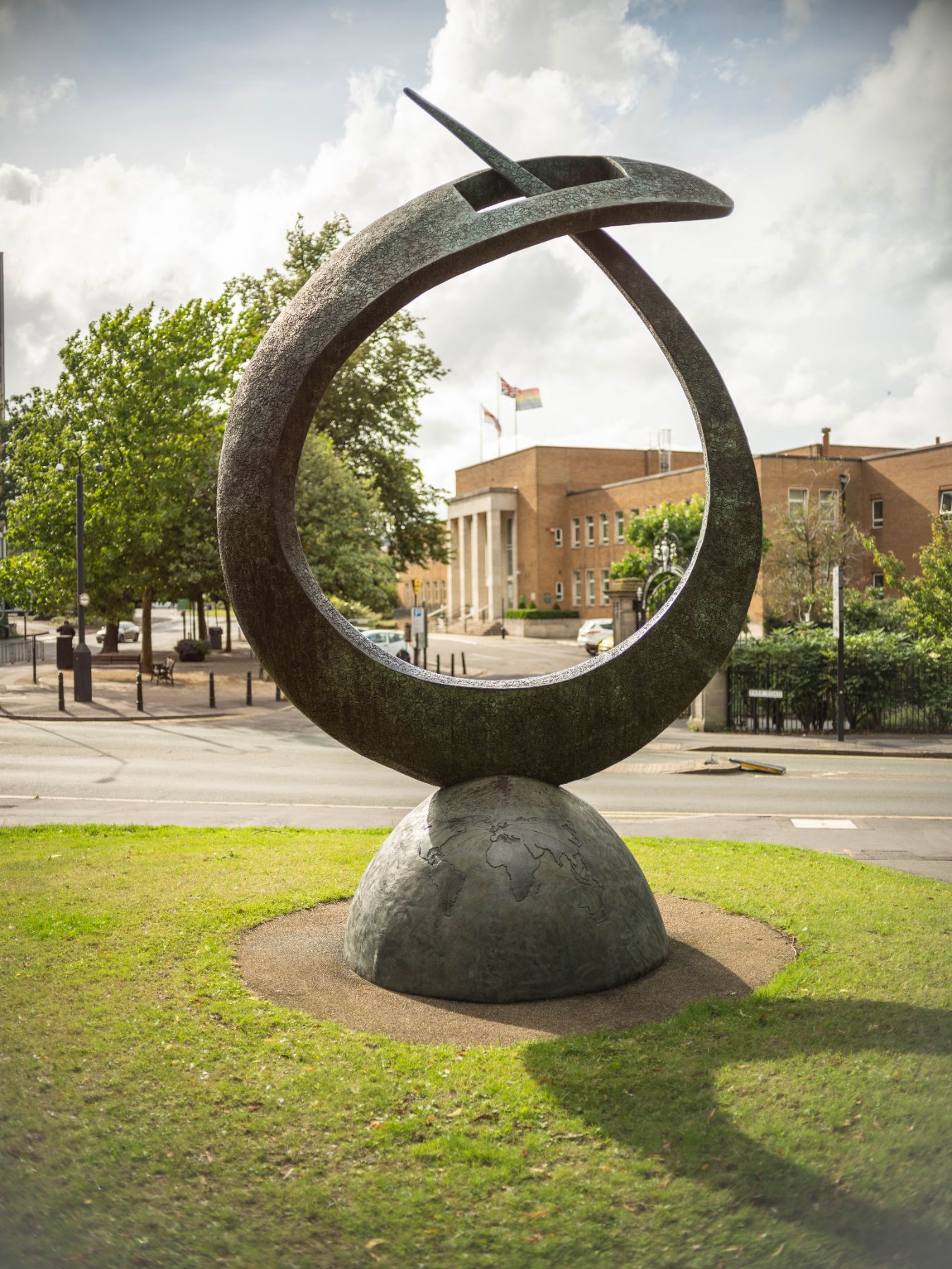 A large bronze ring sculpture balanced on a half-globe stands on a grass verge, with Rugby Town Hall and its flagpoles visible through the ring.